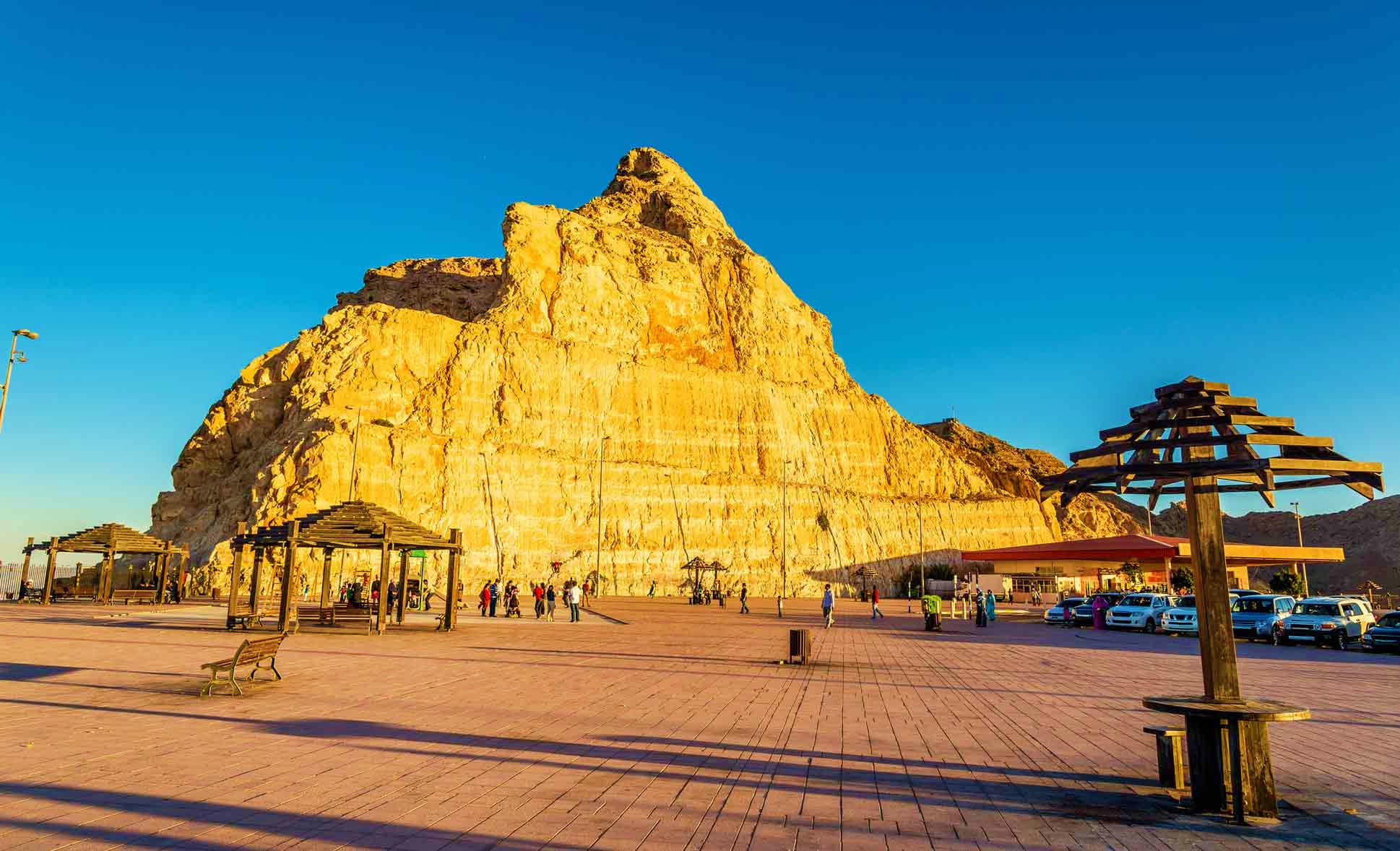 Golden limestone mountain rising above a waterfront promenade with wooden pergolas and tourists in Muscat, Oman
